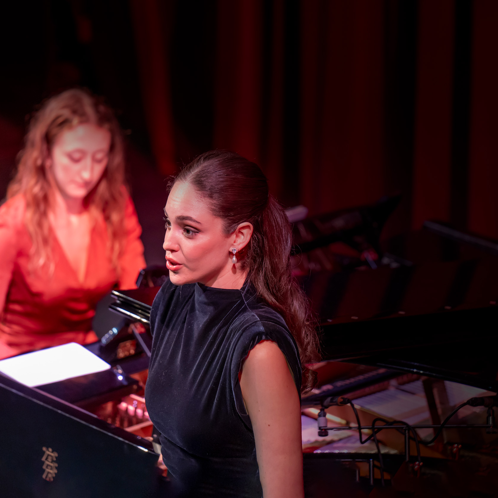 "Your time to shine". A singer wearing a black dress, performing on stage, with a pianist accompanying them on stage in an orange dress.